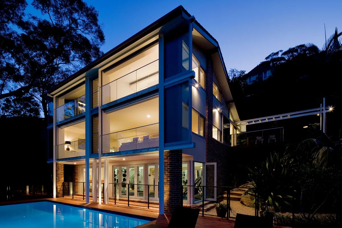 Modern two-story home with glass walls, warm interior lighting, and a swimming pool reflecting blue evening light to the front.