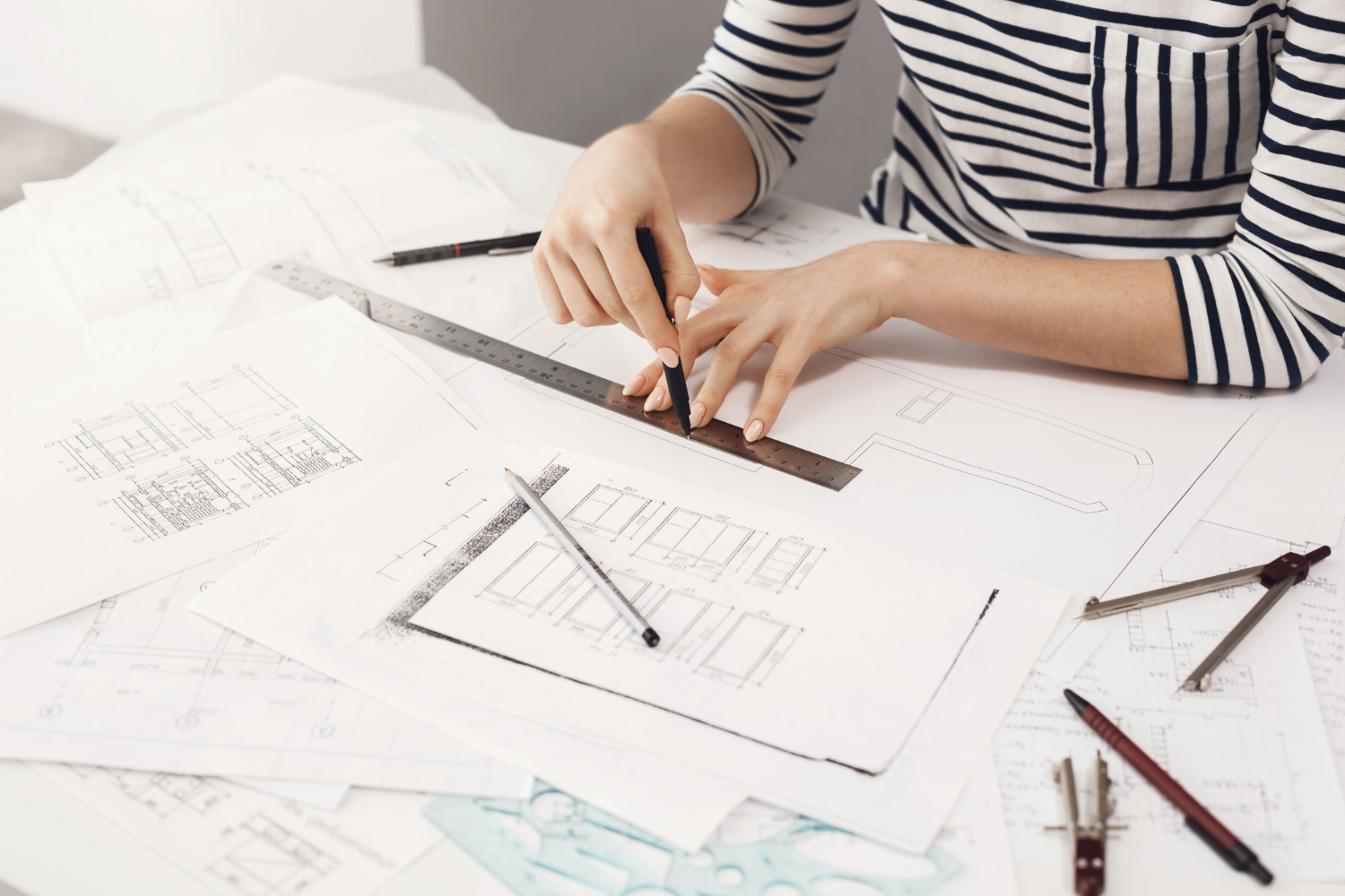 Designer drafting architectural plans with a ruler and pencils on white blueprint sheets on a desk.