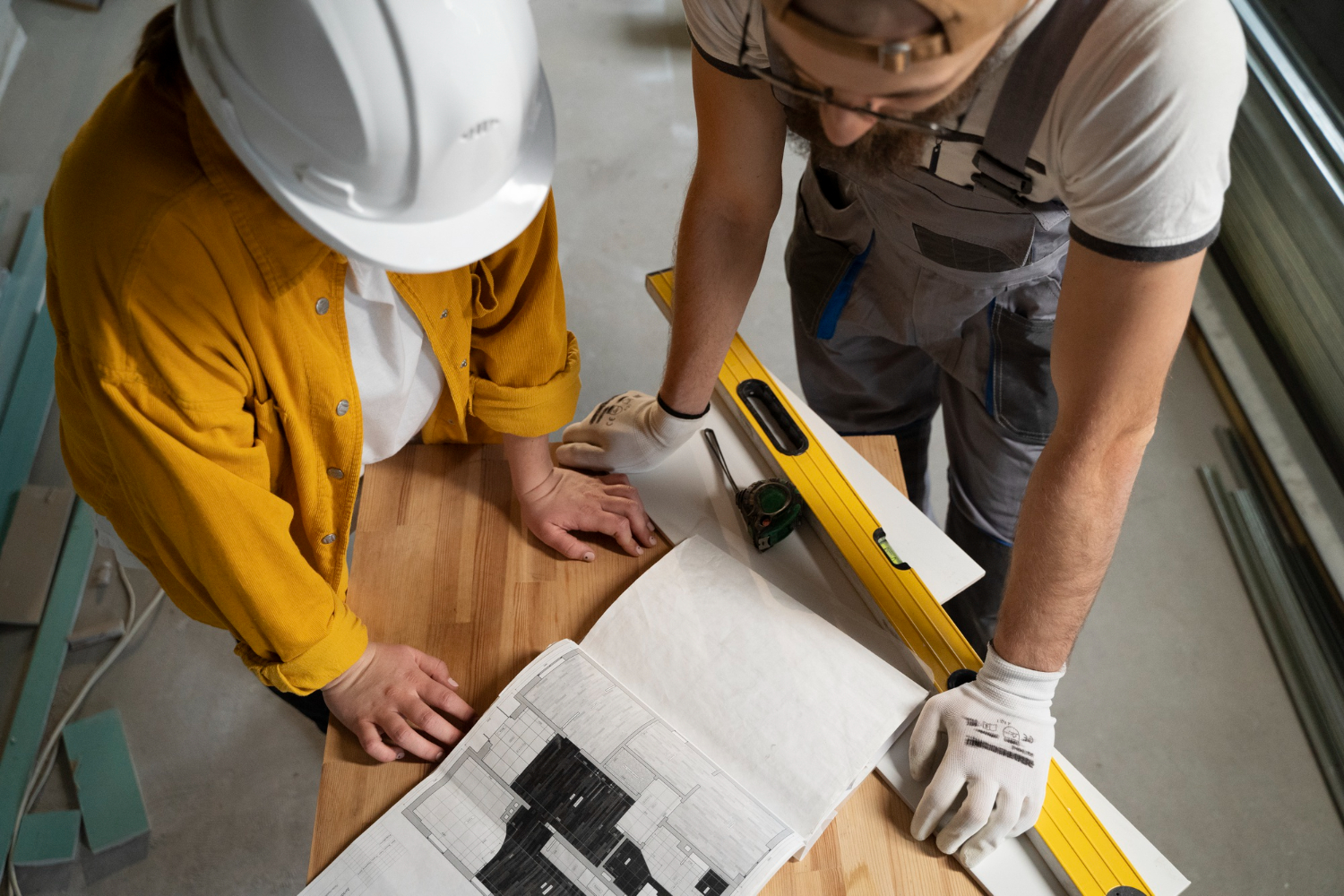 Two construction workers in safety gear measure a wooden board on a workbench using a level, with blueprints spread nearby.